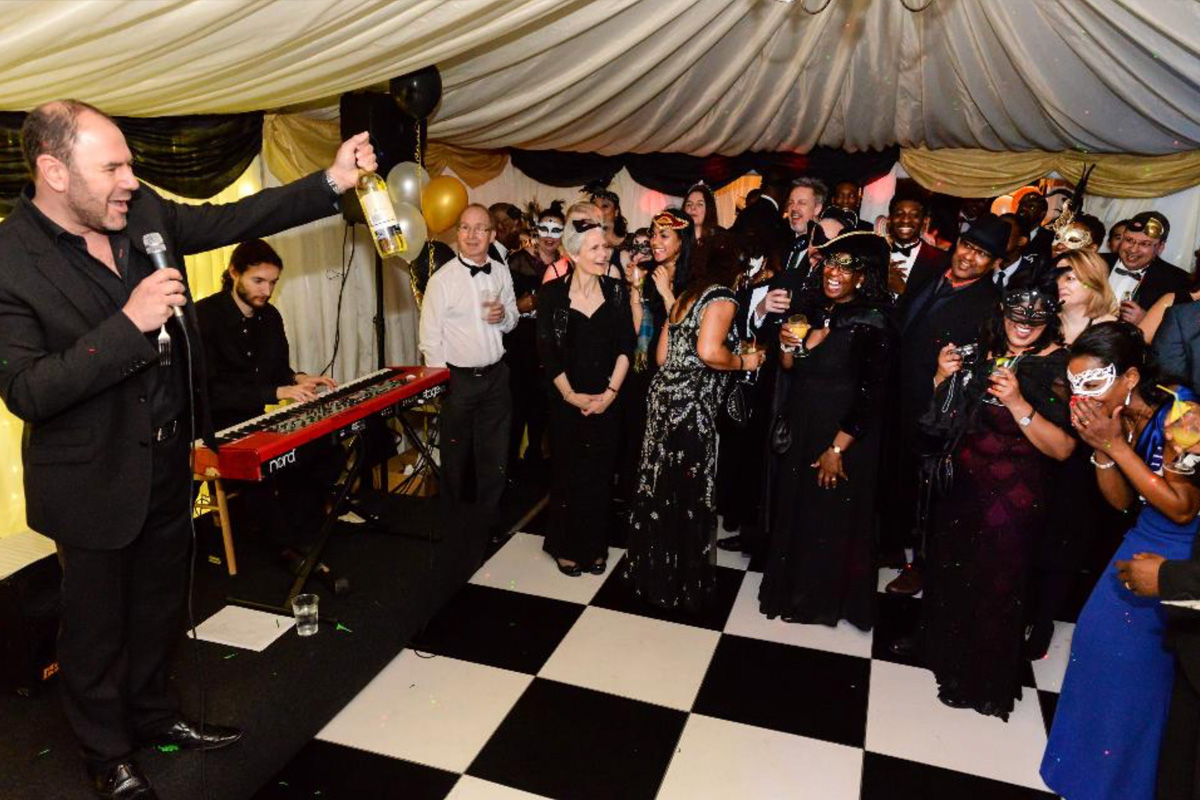 Man with microphone and bottle on stage beside keyboard player at masked party with black and white checkered floor.