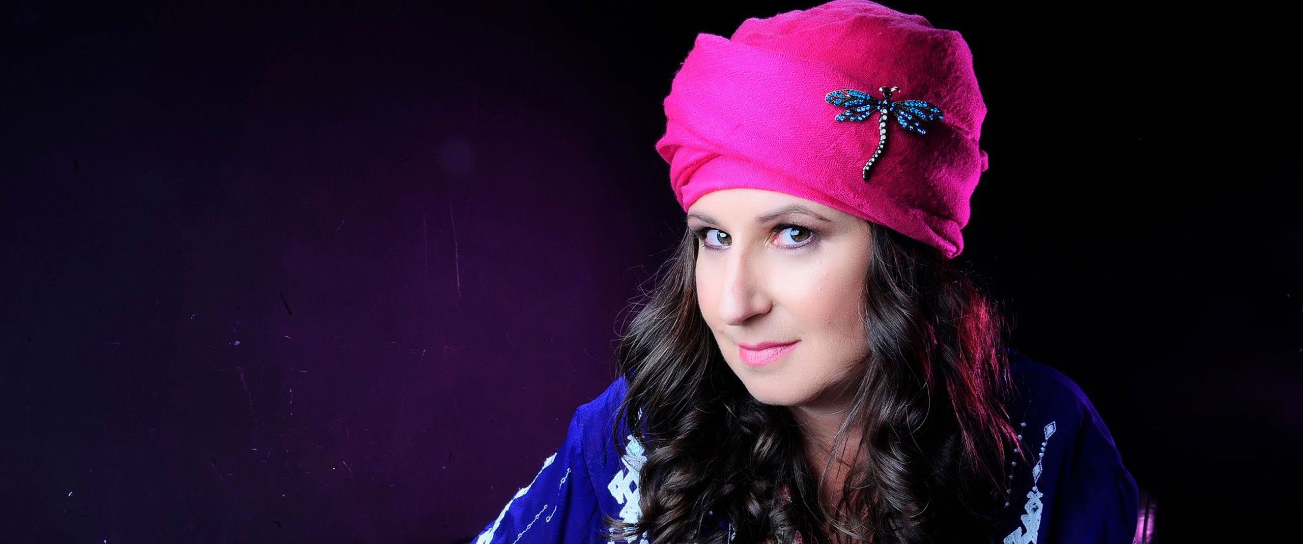 Woman with curled hair wearing a bright pink hat with a blue dragonfly pin, smiling slightly.